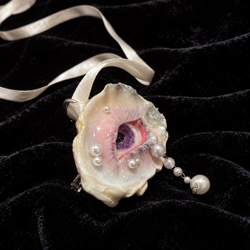 Angled view of Nyra Lavender Gaze oyster-eye jewelry with a pale ribbon and pearl-like drop.